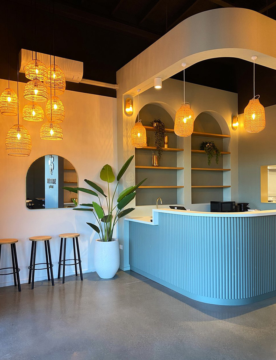 IMG_8305 Elegant restaurant reception with a curved grey counter, warm pendant lights, arched shelving, and a potted plant